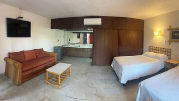 A hotel room with a sofa, a small table, two beds, a TV, a mirror, and a kitchenette.