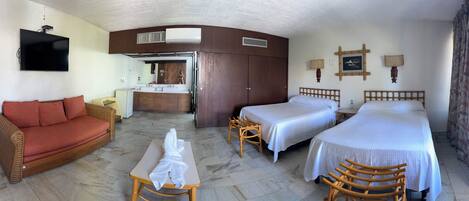 Habitación de hotel con sofá, dos camas, una mesa con una toalla, baño con espejo y televisión.