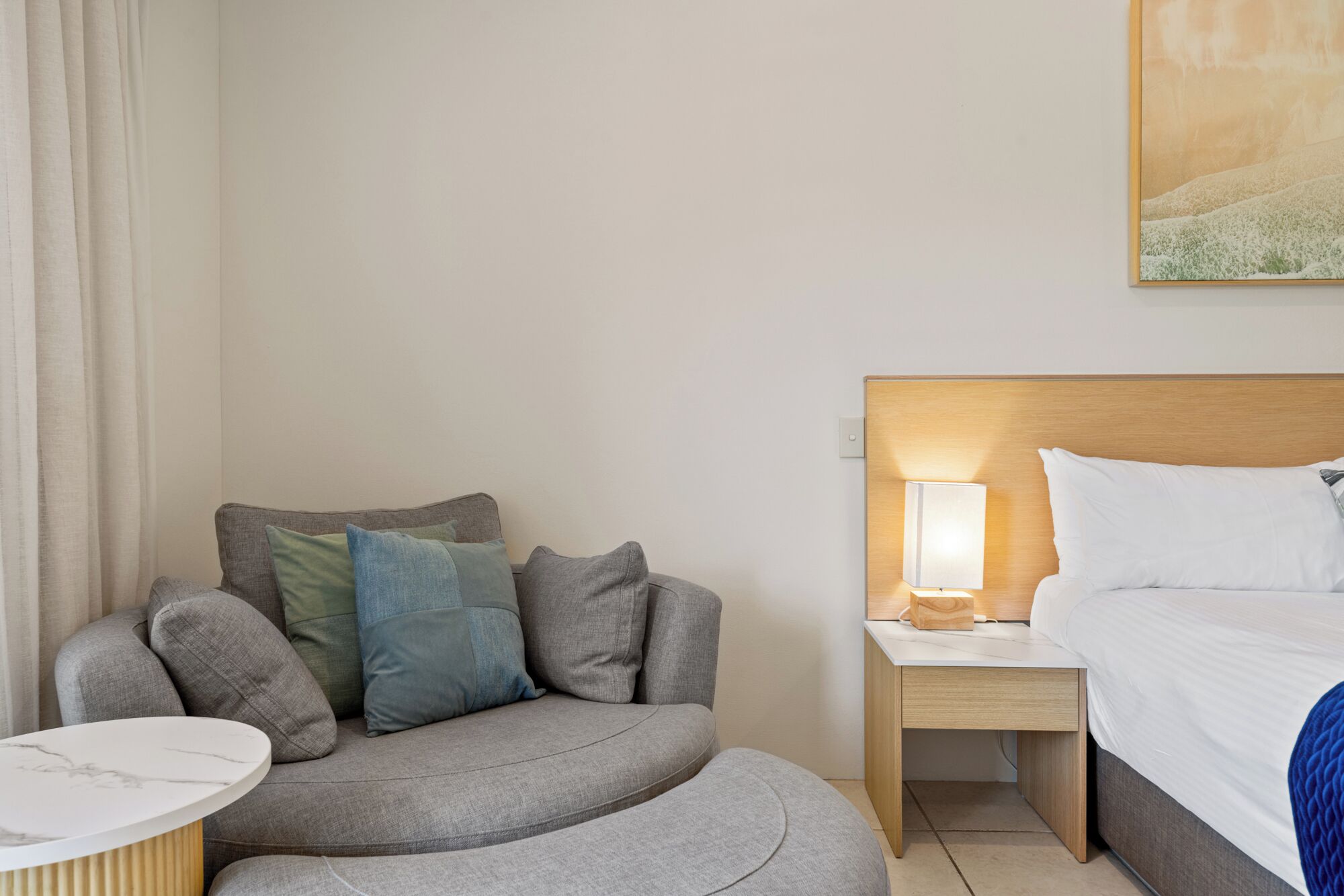 A hotel room with a bed, a nightstand with a lamp, a sofa with cushions, and a small table.