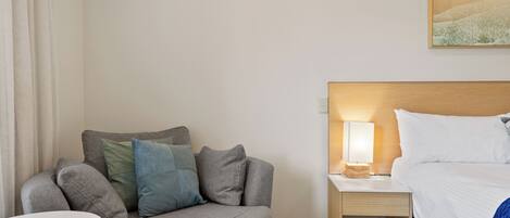 A hotel room with a bed, a nightstand with a lamp, a sofa with cushions, and a small table.