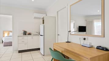 A hotel room with a kitchenette, a bed, a desk with a chair, a mirror, a TV, and a bedside table with a lamp and a phone.