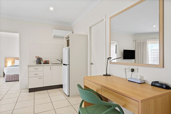 A hotel room with a kitchenette, a bed, a desk with a chair, a mirror, a TV, and a bedside table with a lamp and a phone. - Terrigal Sails Serviced Apartments (Terrigal)