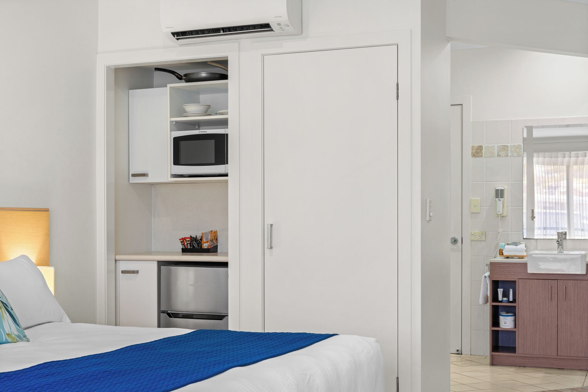 A hotel room with a bed, a microwave, a mini-fridge, and a bathroom with a sink and mirror.