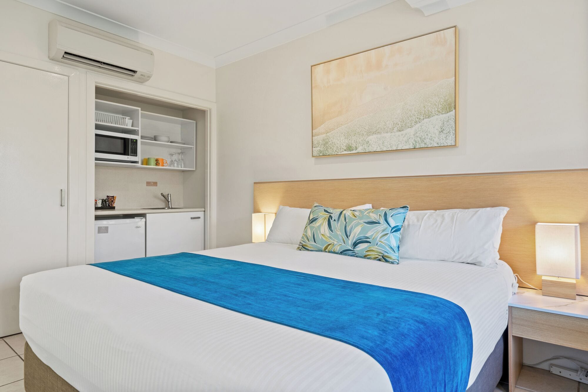 A hotel room with a bed, a nightstand, a desk, a kitchenette, and a painting on the wall.