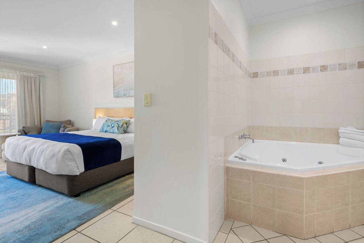 A hotel room with a bed, a blue rug, a bathroom with a bathtub, and a window with curtains.
