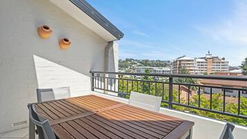 Een balkon met een houten tafel en stoelen, twee terracotta potten aan de muur, en uitzicht op gebouwen en groen.