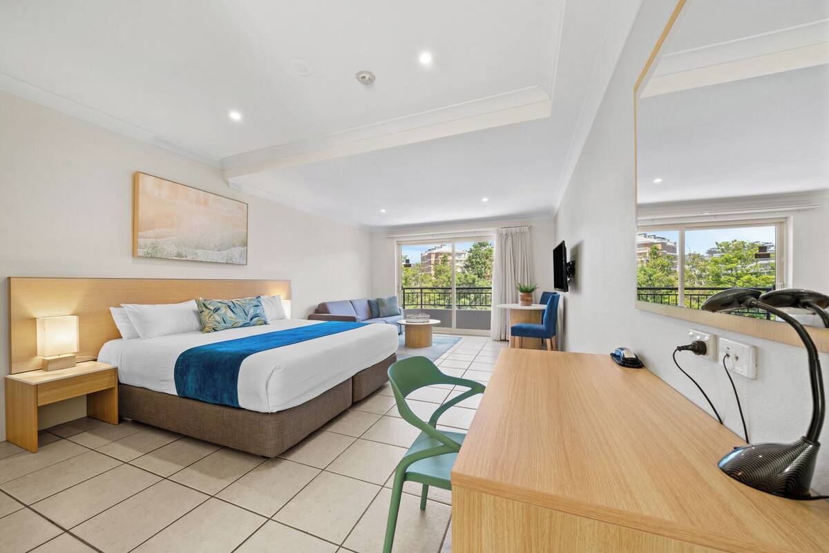 A modern hotel room with a bed, desk, chair, and a view of the outdoors.