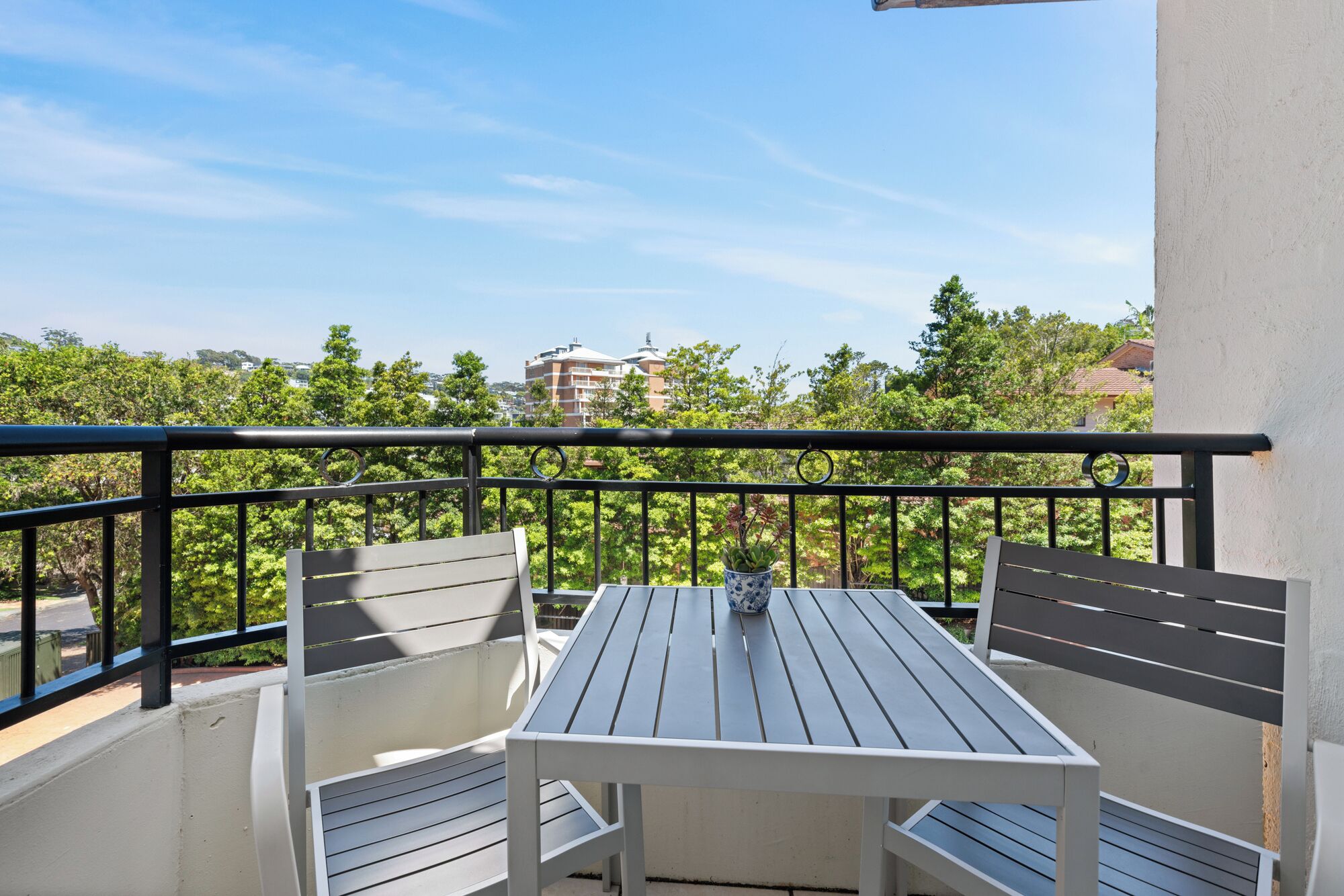 A balcony with a table and two chairs, a potted plant, and a view of buildings and trees.