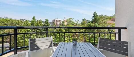 A balcony with a table and two chairs, a potted plant, and a view of buildings and trees.