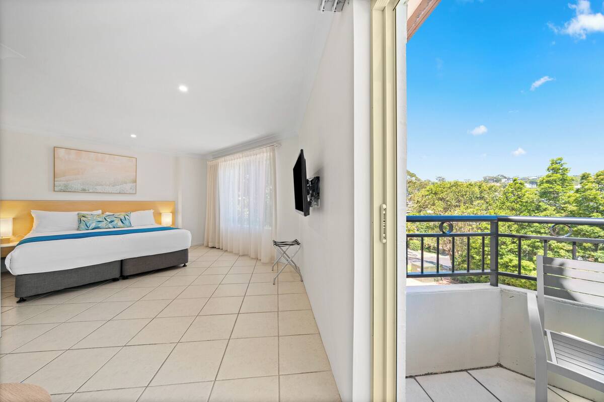 A hotel room with a bed, a TV mounted on the wall, and a balcony with a view of greenery.