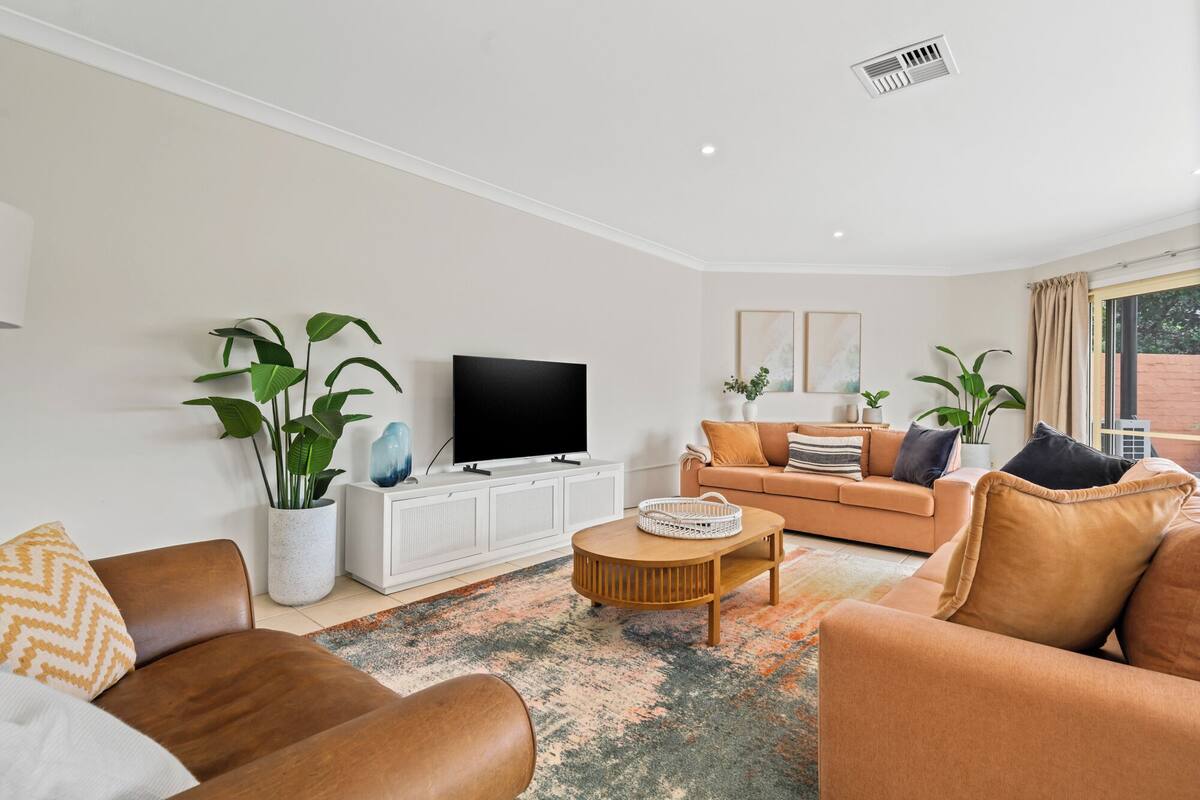 A modern living room with a sofa, a coffee table, and a TV on a white cabinet. There are plants and a window with curtains.