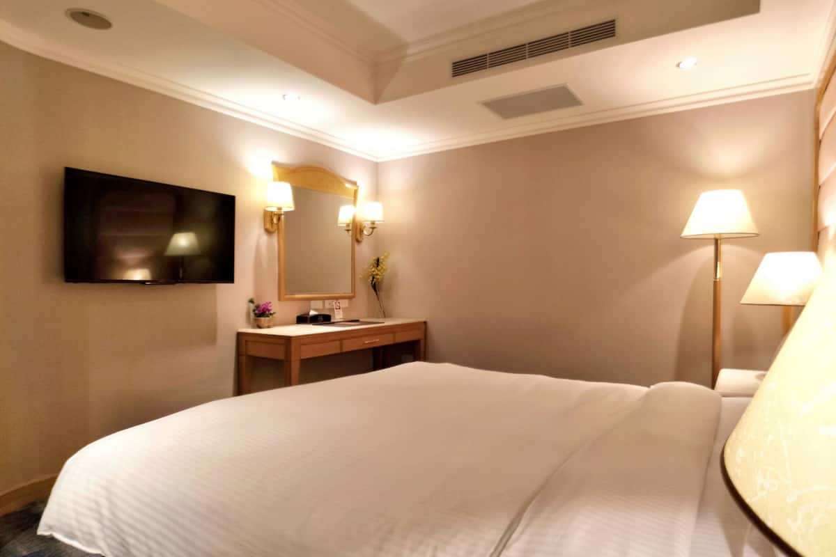 A hotel room with a large bed, a wooden vanity with a mirror, a flat-screen TV mounted on the wall, and a standing lamp.