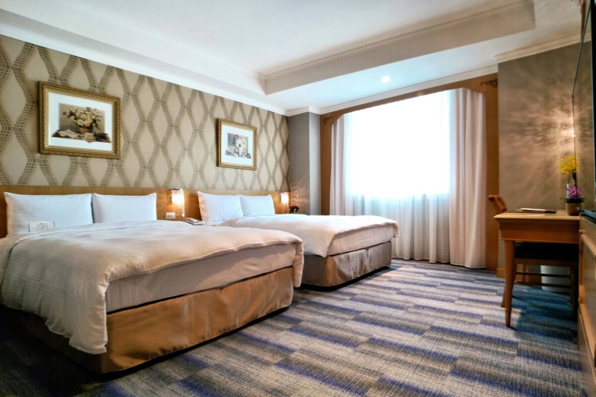 A hotel room with two beds, a desk, and a large window.