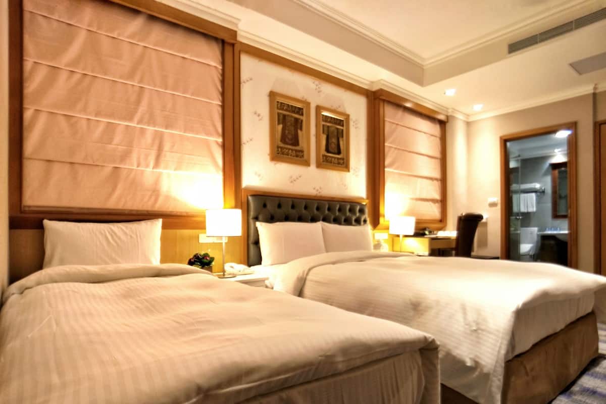 A hotel room with two beds, a large window with blinds, a headboard with a tufted design, and a framed picture on the wall.