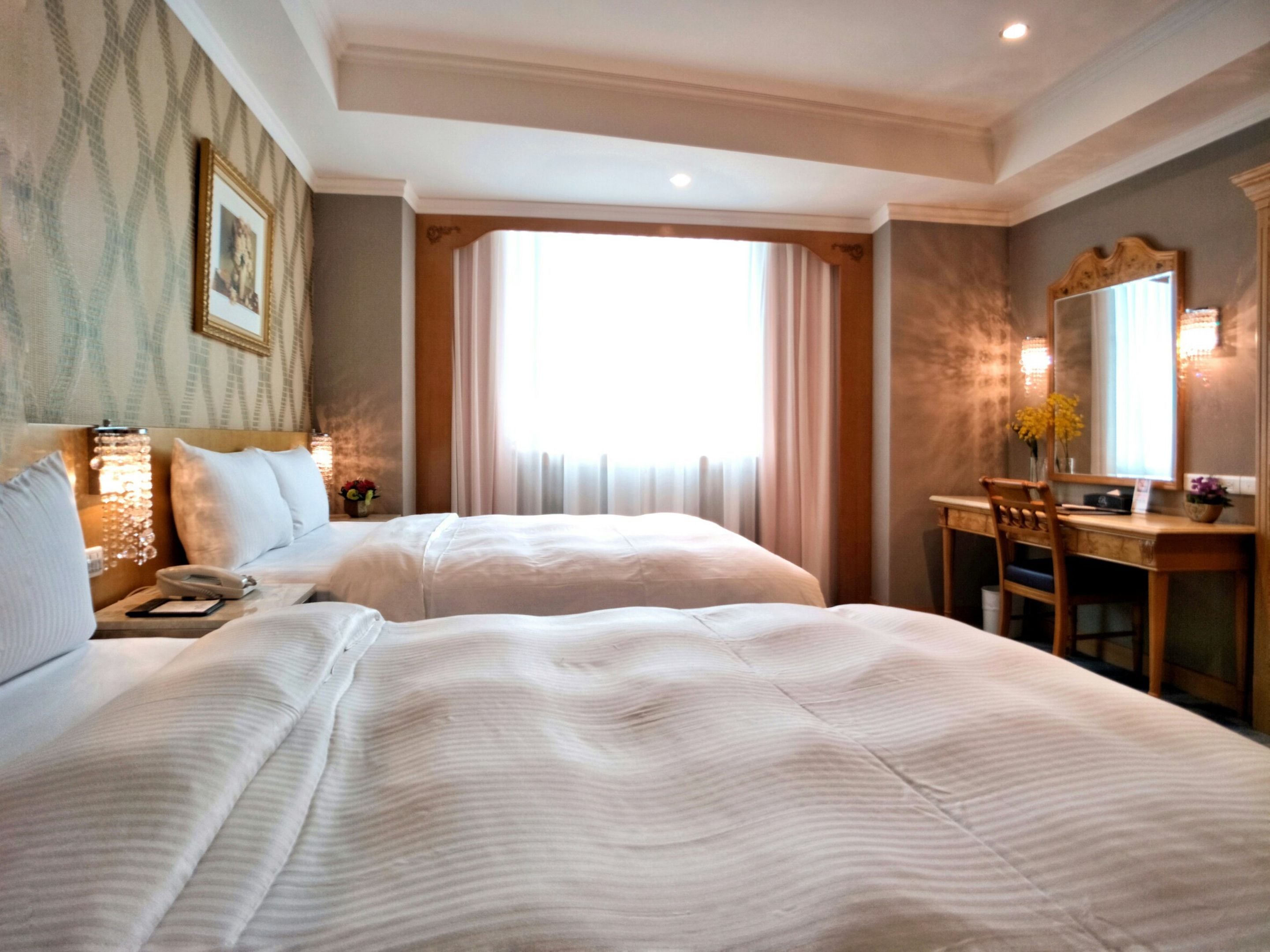 A hotel room with two beds, a desk, a chair, a mirror, and a window with curtains.