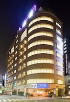 Un hotel de varios pisos con un llamativo letrero que dice “HOTEL” al nivel de la calle, que presenta un diseño arquitectónico moderno con fachada curva y múltiples ventanales.