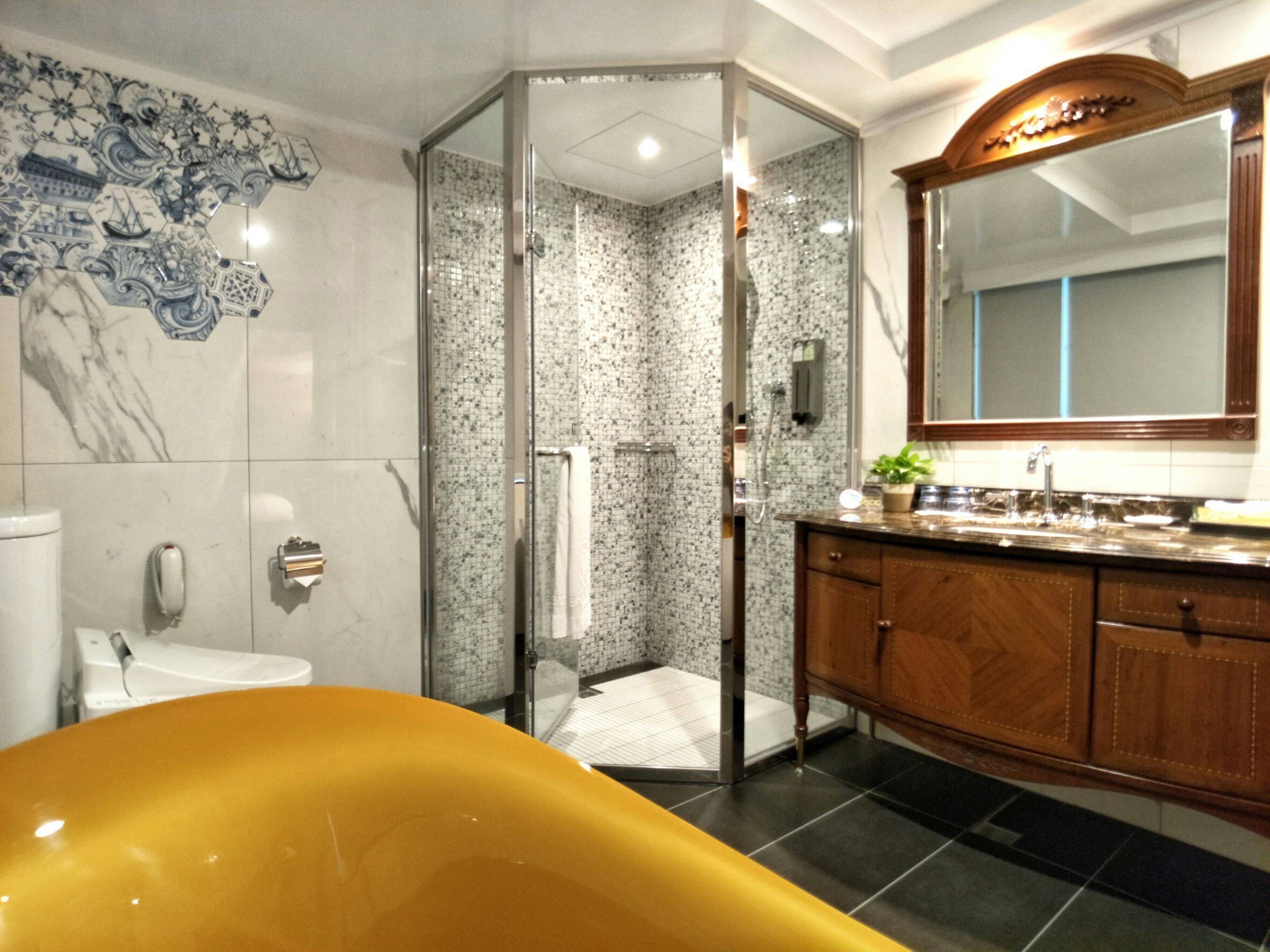 Une salle de bain avec une baignoire jaune, un espace de douche en verre, un mur recouvert de carreaux de marbre, un meuble-lavabo en bois et un grand miroir.