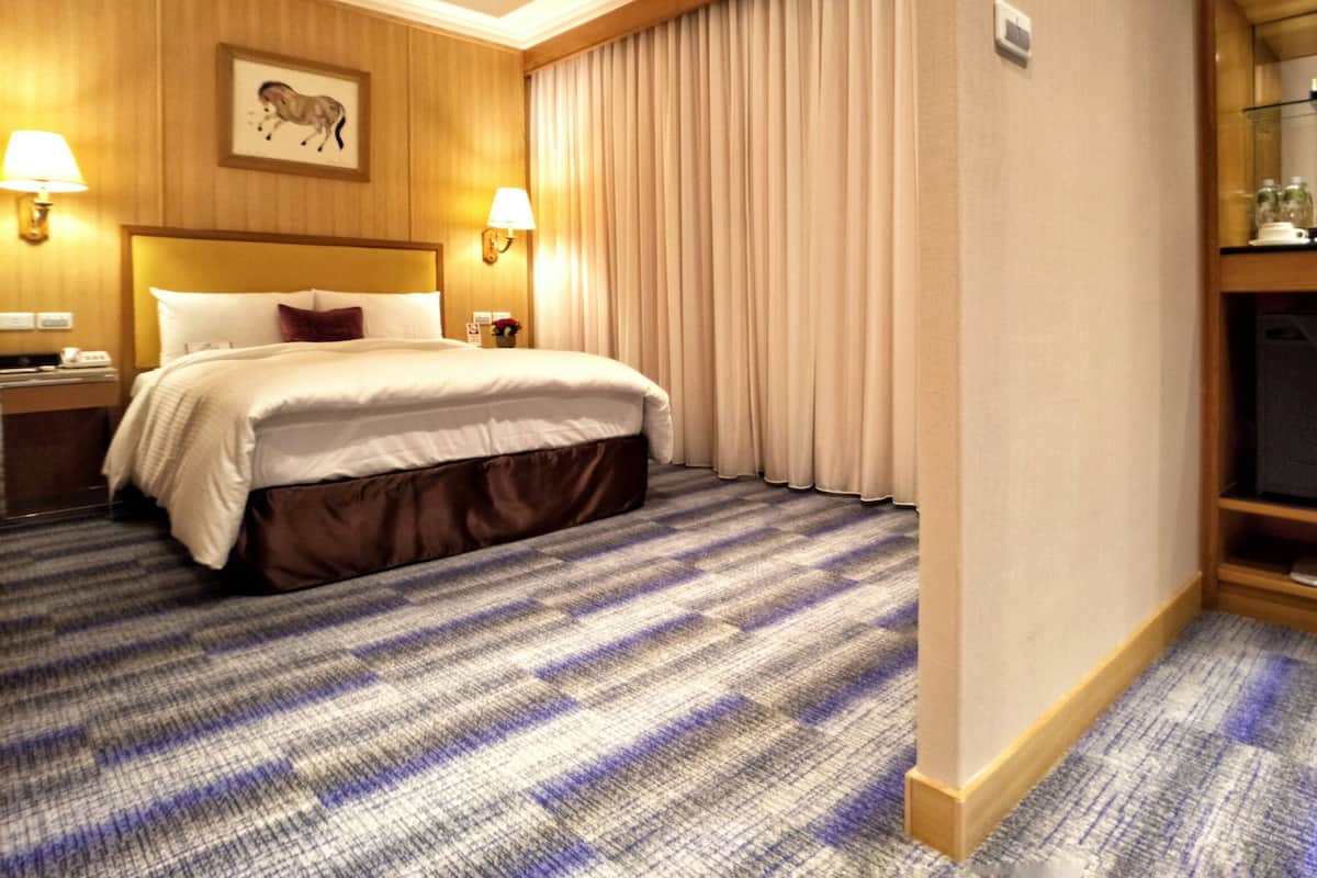 A hotel room with a large bed, two bedside lamps, a headboard with a mounted picture, a nightstand, a carpeted floor, and a window with curtains.