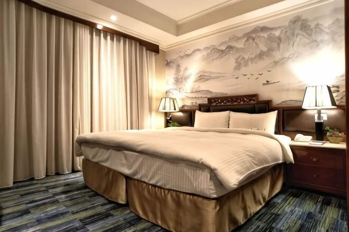 A hotel room with a large bed, curtains, a nightstand with a lamp, and a wall with a painting.