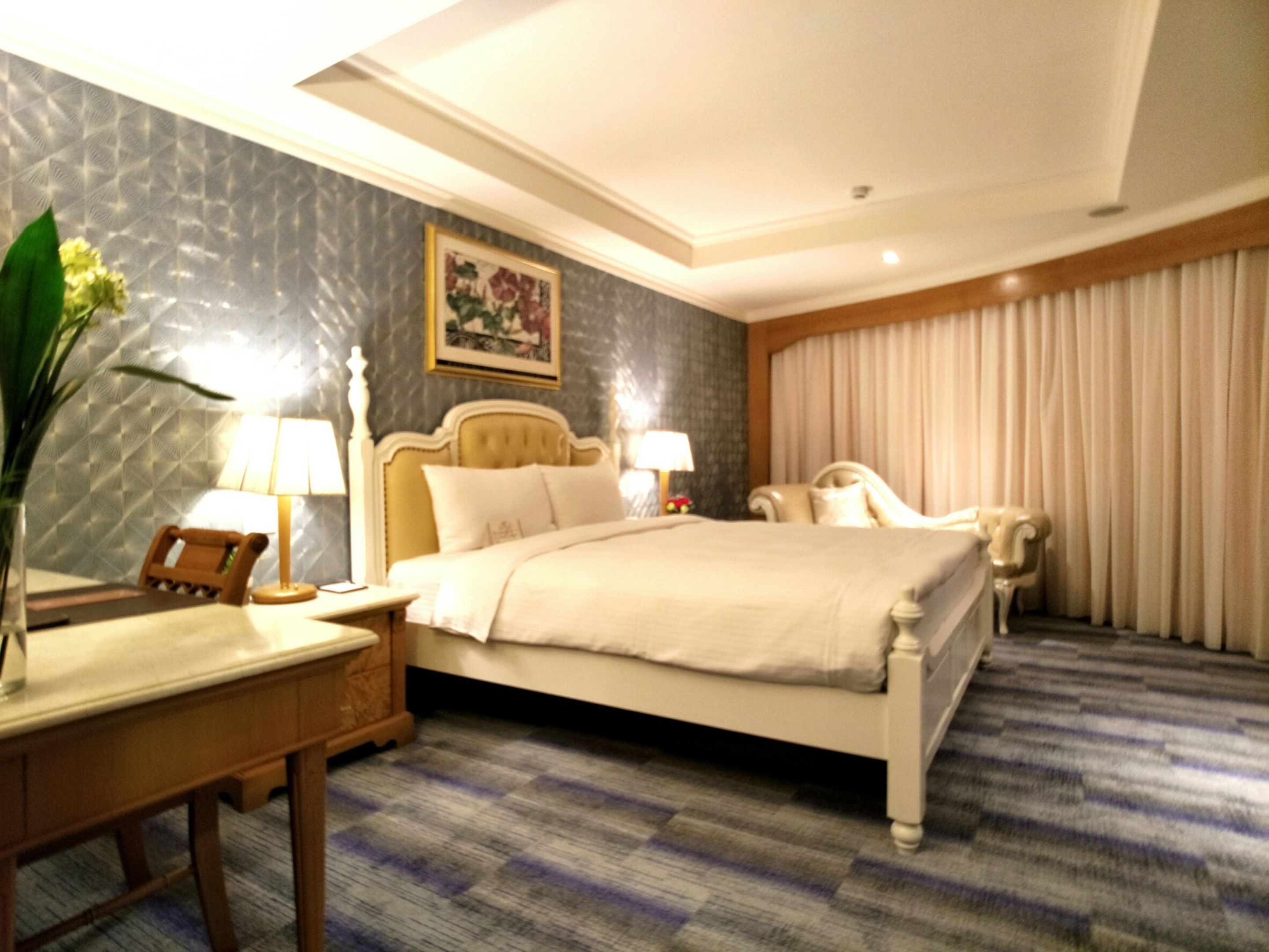 Une chambre d’hôtel avec un grand lit, deux tables de chevet, un bureau, une chaise, une lampe, un vase avec des fleurs et un tableau au mur.