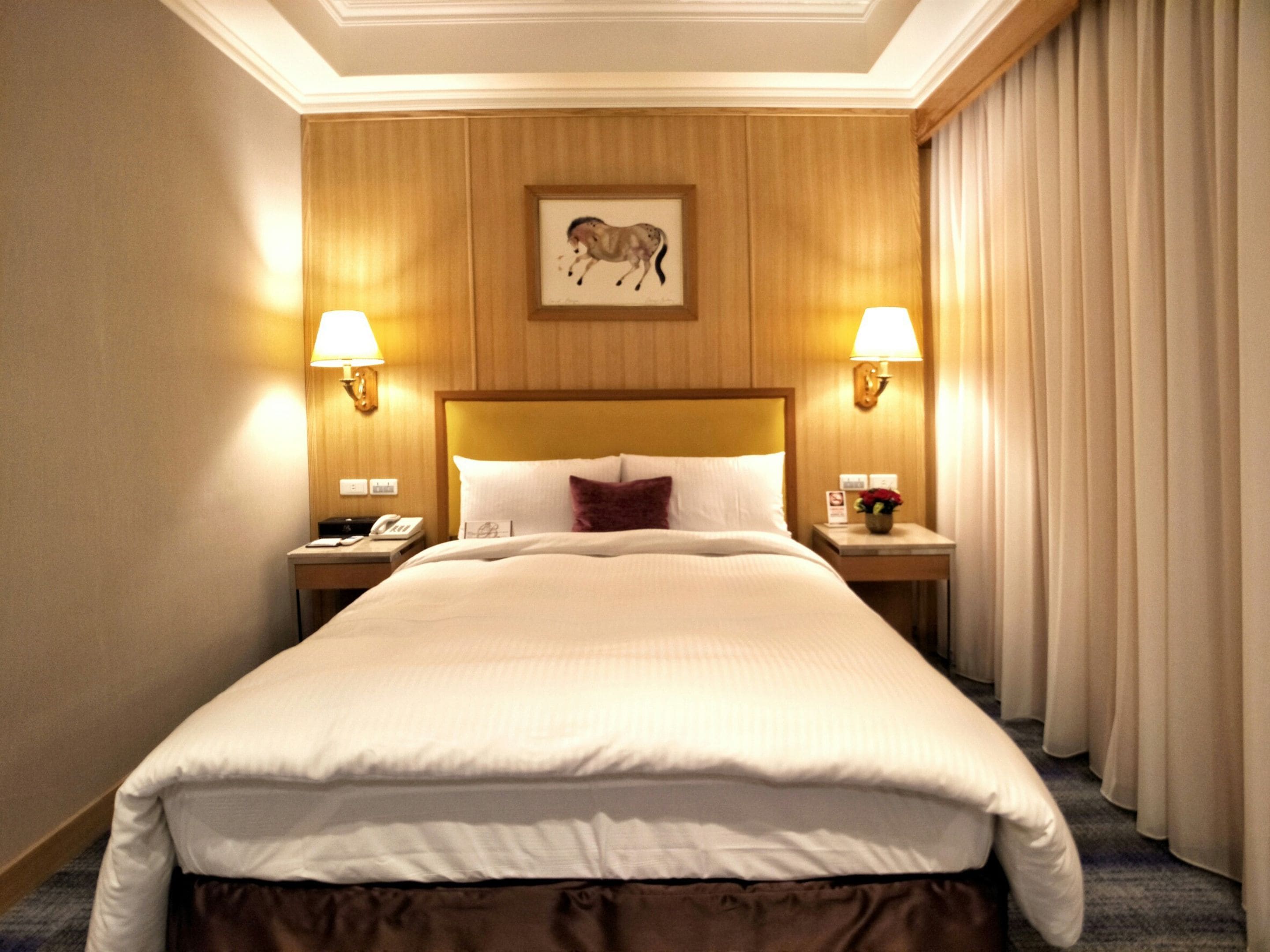 A hotel room with a large bed, two bedside tables with lamps, a mounted picture of a horse, and a view of the outside curtains.
