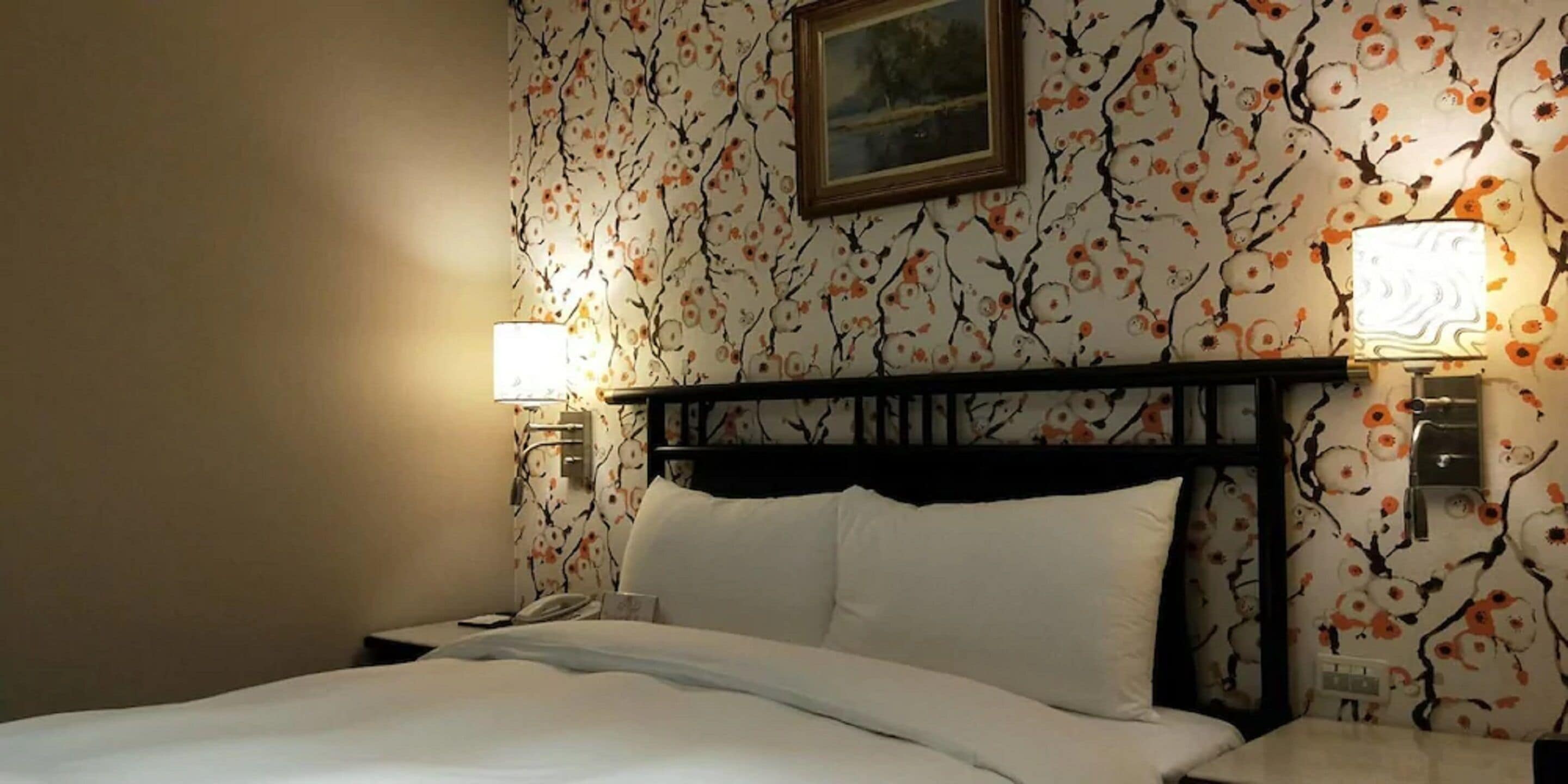 A hotel room with a bed, two bedside lamps, a nightstand, and a floral wallpaper pattern.