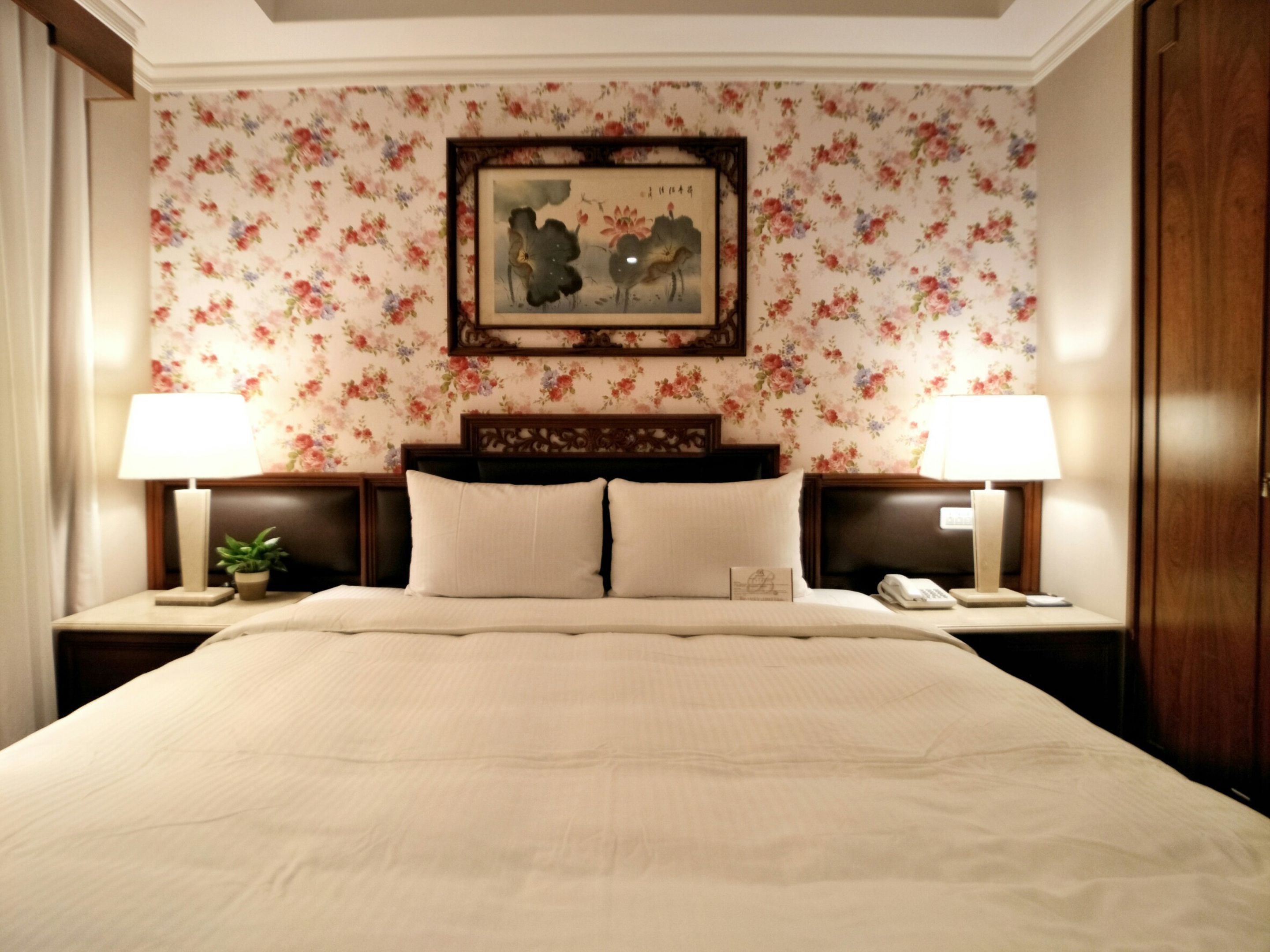 A neatly made bed with a headboard, two bedside tables with lamps, a floral wallpaper, and a framed artwork above the bed.