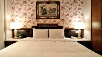 A neatly made bed with a headboard, two bedside tables with lamps, a floral wallpaper, and a framed artwork above the bed.
