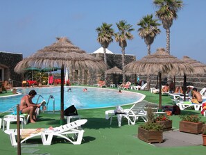 Zona de piscina con sillones de descanso, sombrillas y palmeras.