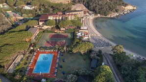 Vista aérea de un hotel costero con varias construcciones, cancha de tenis y playa con sombrillas.