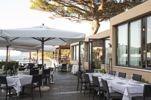 Un área de comedor al aire libre con mesas dispuestas para dos, sombrillas y vista al mar.