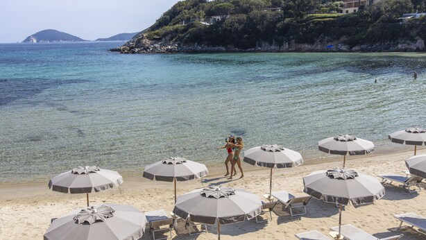 Una playa con sombrillas y tumbonas, vista despejada del mar y una colina con edificios.
