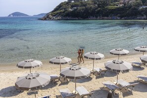 Una playa con sombrillas y tumbonas, vista despejada del mar y una colina con edificios.
