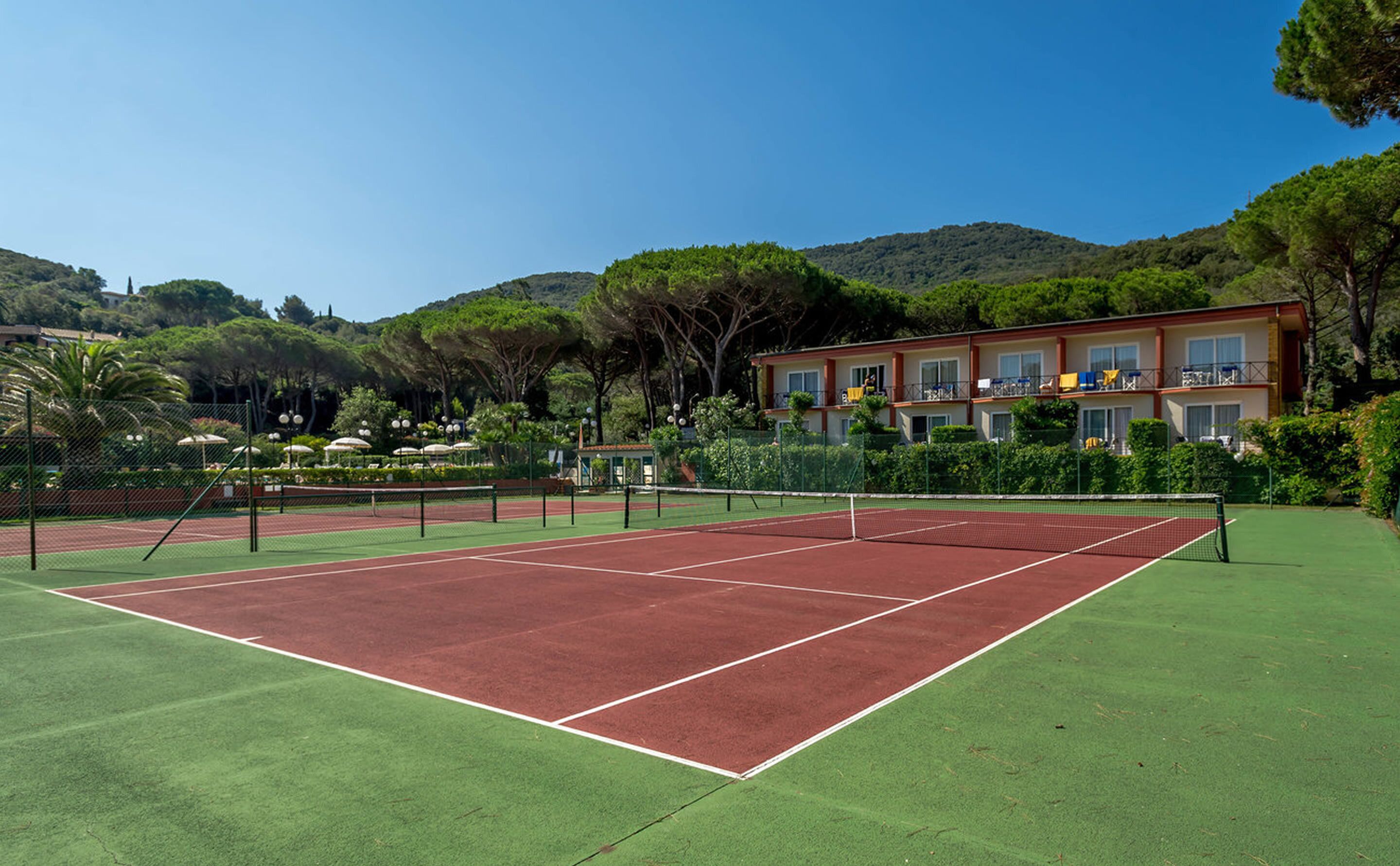A tennis court with a net, surrounded by greenery and a building with balconies.