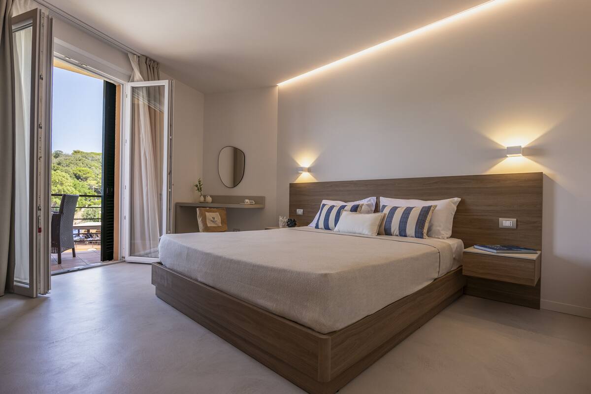 A modern bedroom with a large bed, bedside tables, and a view of greenery through an open door.