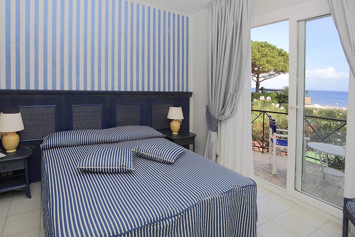 A hotel room with a bed, two bedside tables with lamps, a view of the sea, and a balcony with chairs.