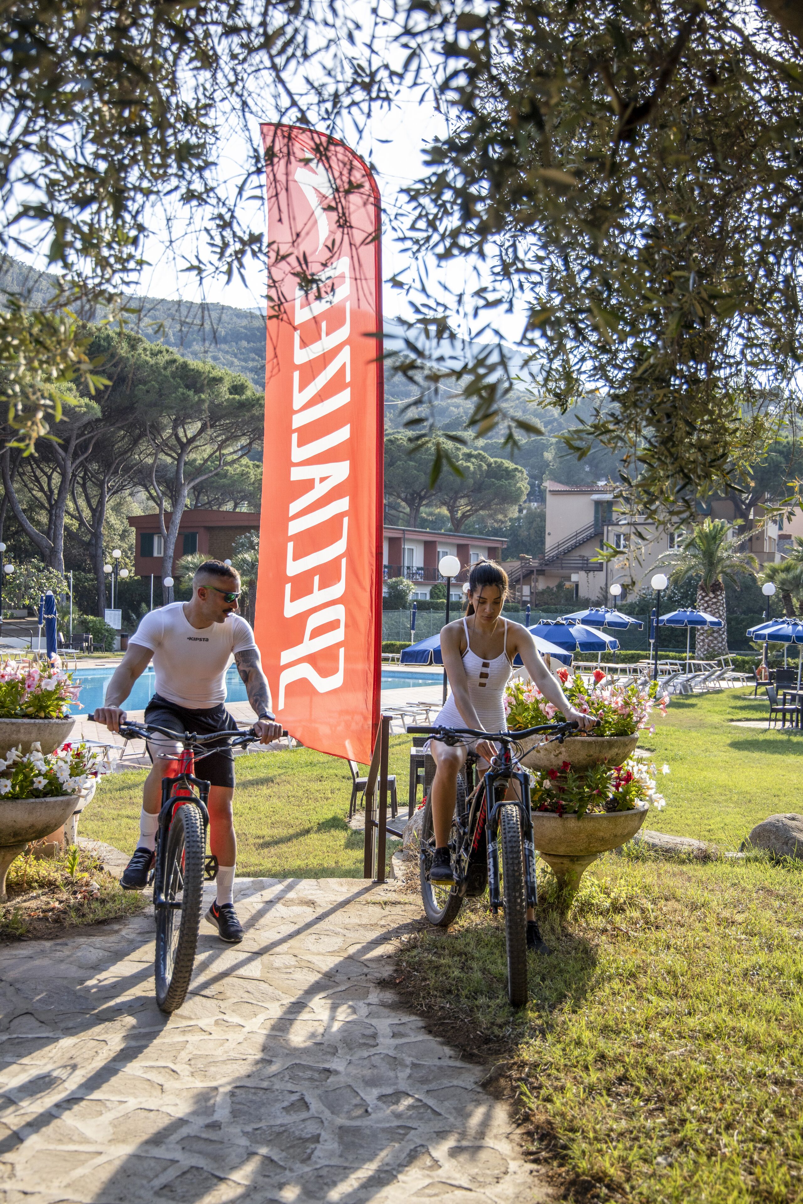 Two people riding bicycles on a stone path with a red banner in the background.