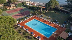 Una gran piscina rodeada de tumbonas y sombrillas, con una cancha de tenis y una zona de playa al fondo.