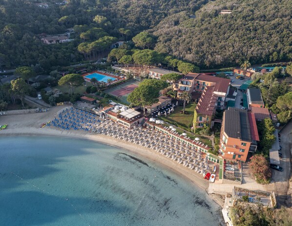 Vista aérea de un complejo turístico frente al mar, con playa, piscina y varios edificios.