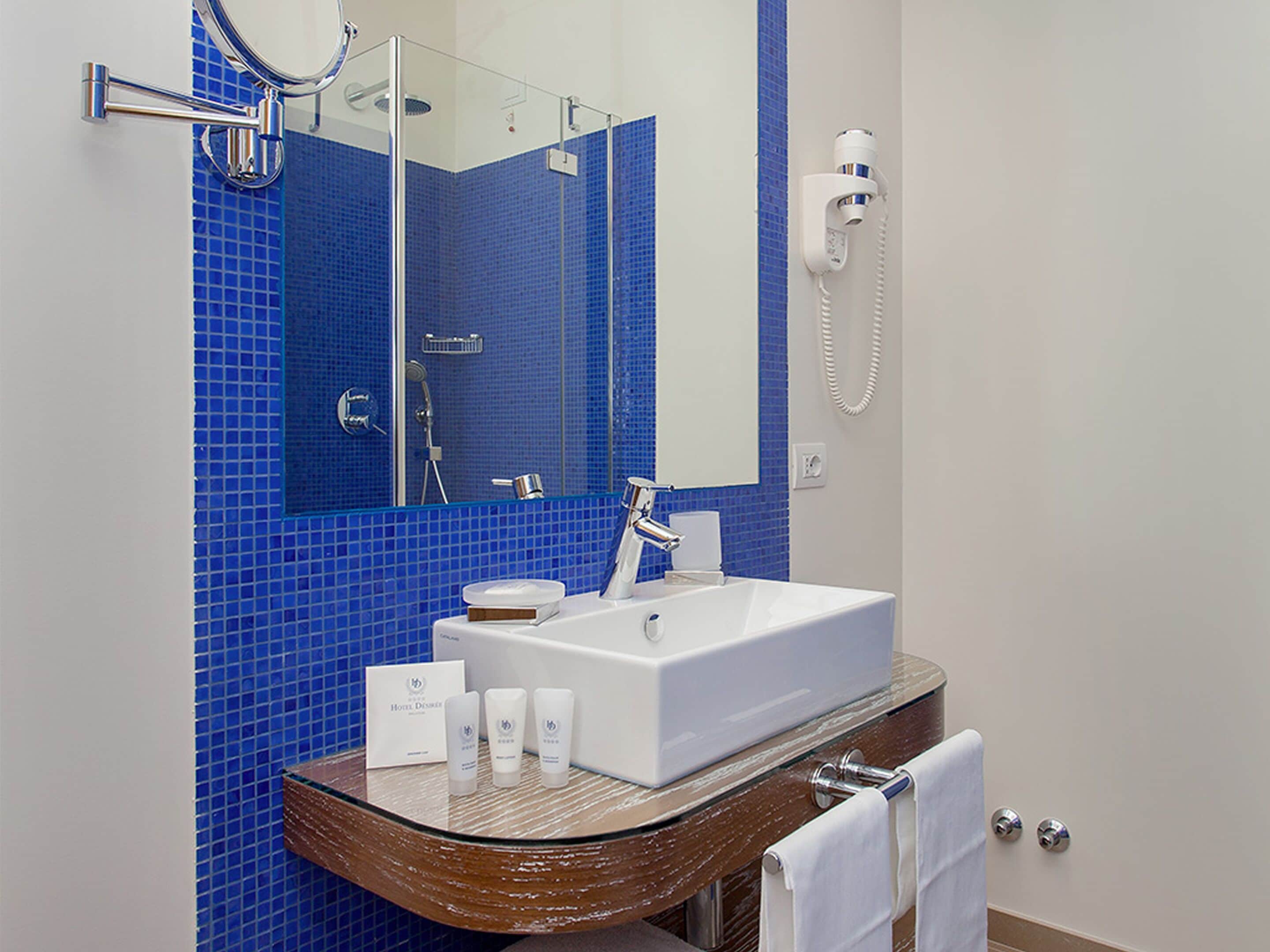 A bathroom with a white sink, blue-tiled walls, a glass-topped vanity, a handheld showerhead, and a towel rack.