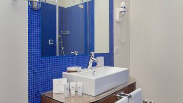 Un bagno con lavandino bianco, pareti rivestite di piastrelle blu, una toeletta con piano in vetro, un soffione a mano e un porta asciugamani.