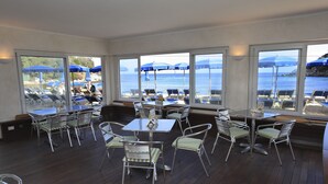 Un área de comedor al aire libre iluminada por el sol, con mesas y sillas, y ventanas amplias con vistas a la playa y el océano.