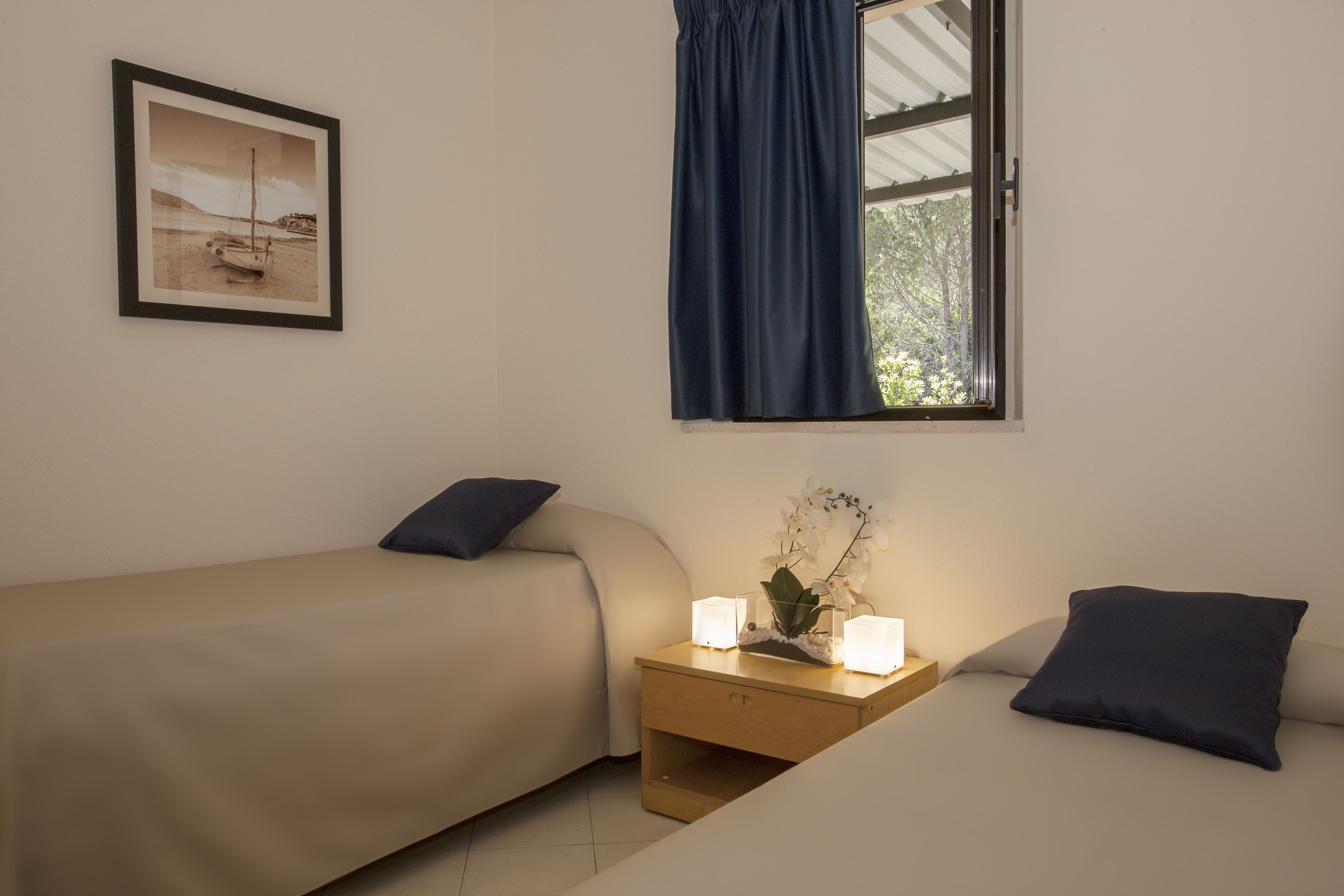 A room with two beds, a nightstand with candles, a framed picture on the wall, and a window with a curtain.