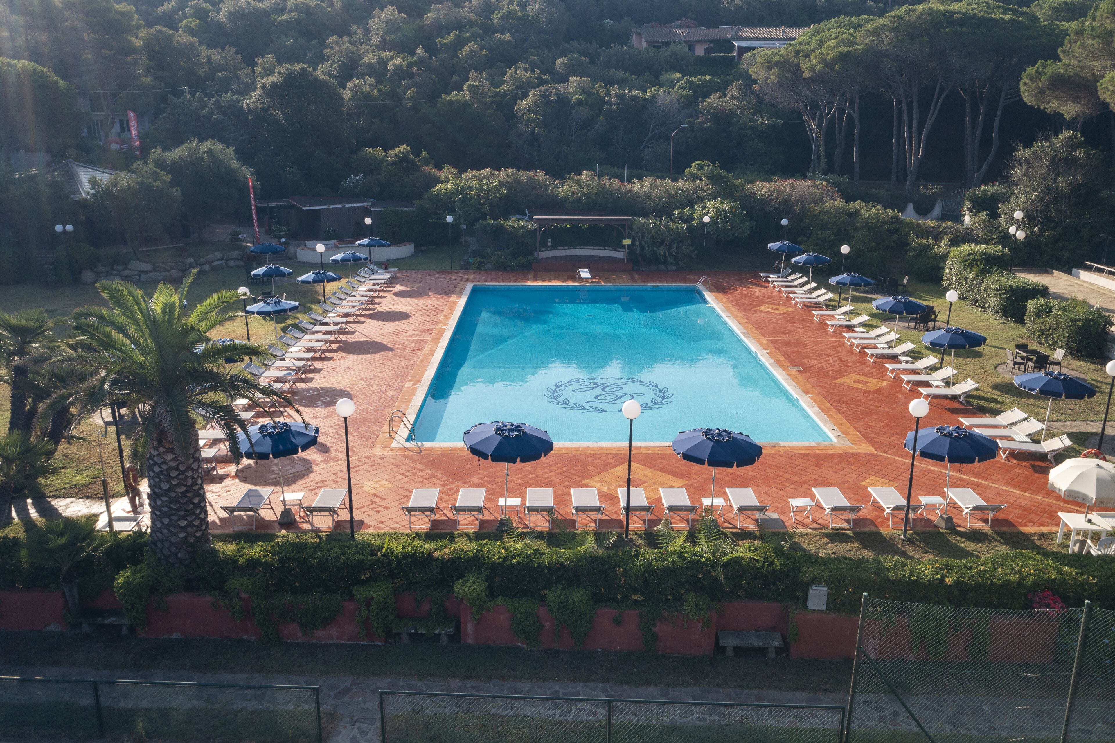 Una piscina rectangular rodeada de sillones de descanso y sombrillas, con una terraza empavedada y vegetación al fondo.