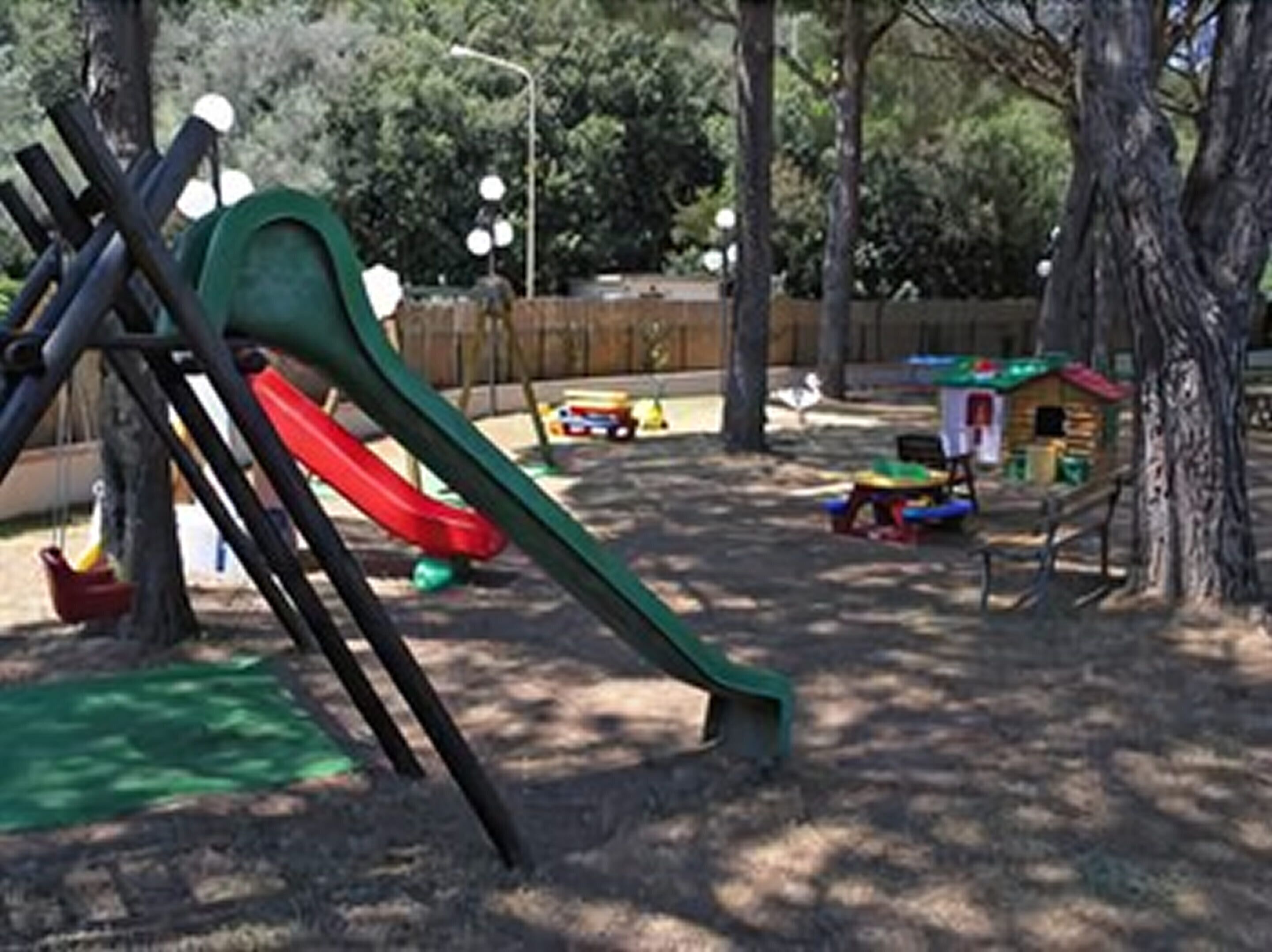 A playground with a green slide and a red slide, surrounded by trees and a wooden fence.