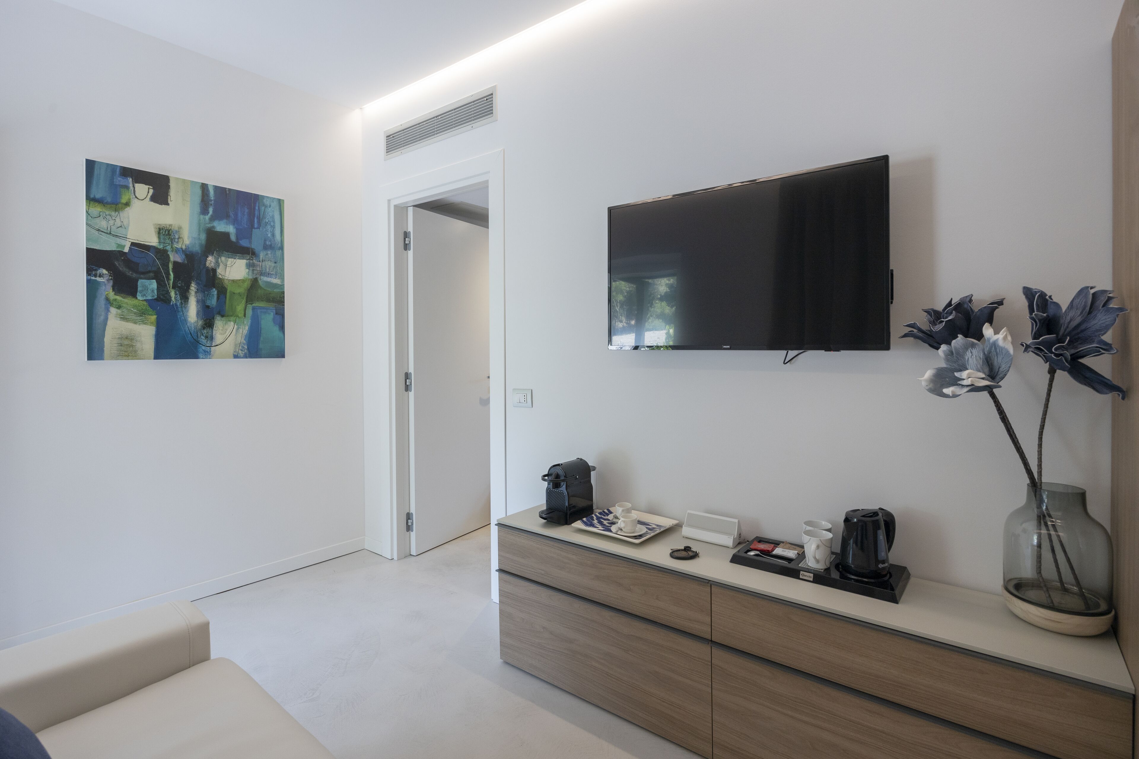 A modern hotel room with a minimalist design, featuring a flat-screen TV, a coffee machine, a vase with flowers, and a painting on the wall.