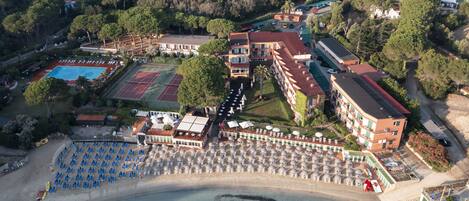 An aerial view of a resort with a swimming pool, tennis courts, and a beach area with umbrellas.