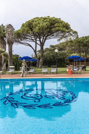 Una piscina con un diseño decorativo, rodeada de sillas de descanso y sombrillas, con árboles y un cielo despejado al fondo.