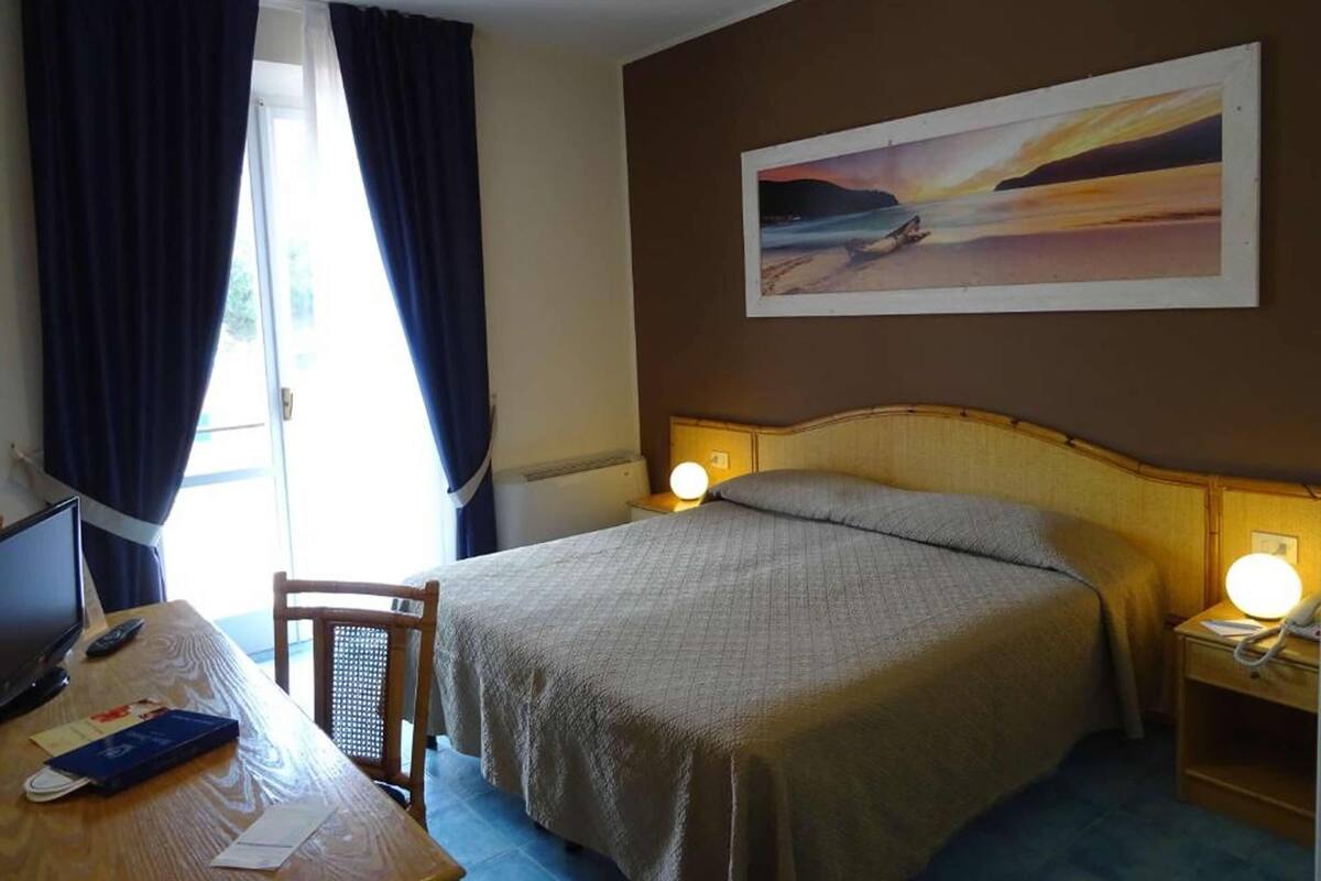 A hotel room with a bed, a desk with a computer, a chair, and a painting on the wall.