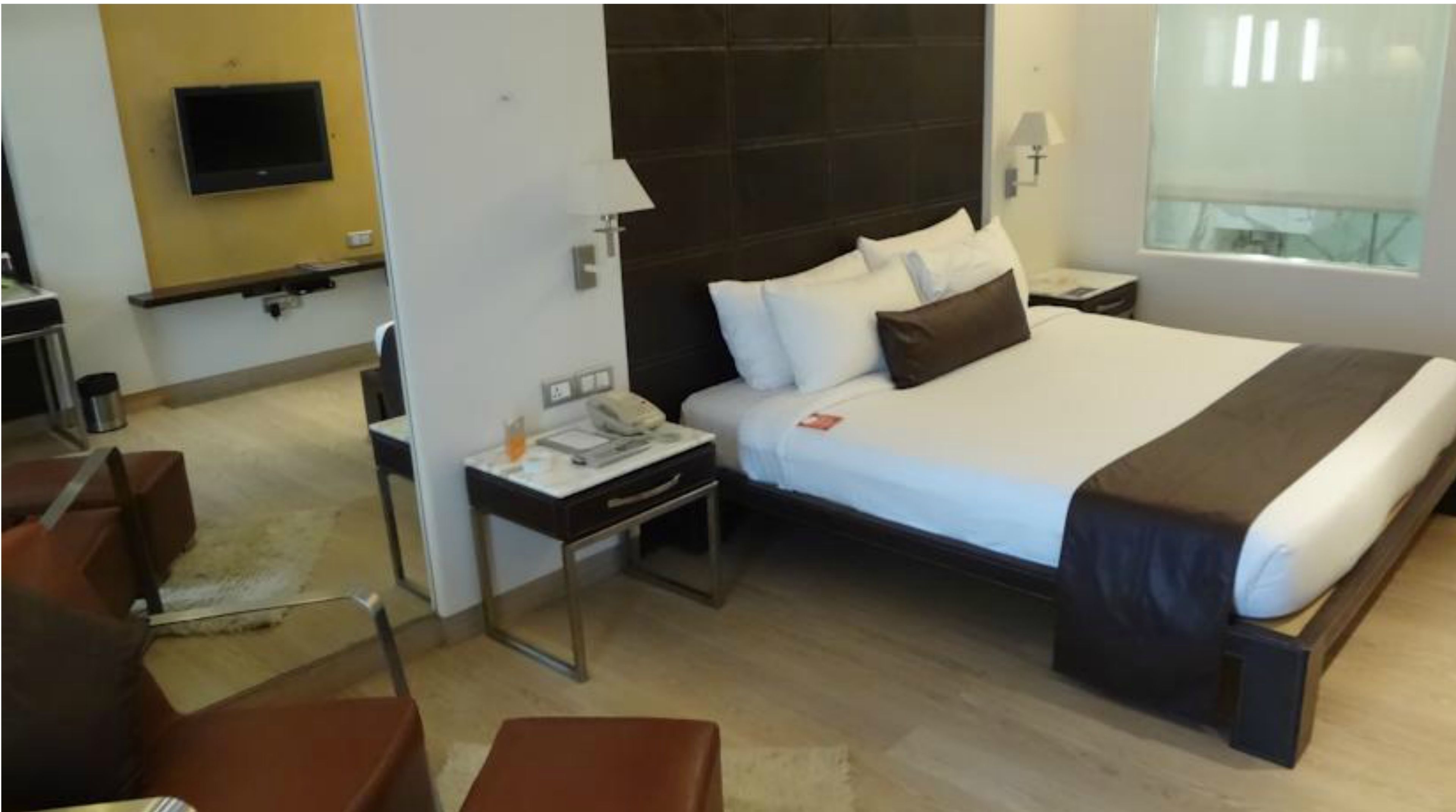 A hotel room with a large bed, a bedside table with a lamp, a TV mounted on the wall, and a window with blinds.